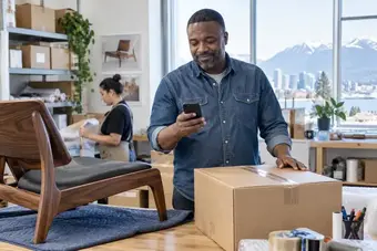 Membre du service à la clientèle vérifiant le statut d’un envoi sur un téléphone, près d’un colis prêt à expédier