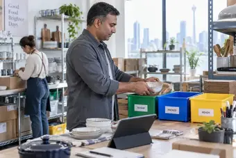 Business owner sorting product orders into labelled, coloured trays beside a tablet